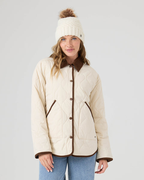 A woman wearing the Roxy Nyvea Quilted Barn Jacket in Fog, along with blue jeans and a cream knit beanie with a brown pompom, stands against a plain light background.