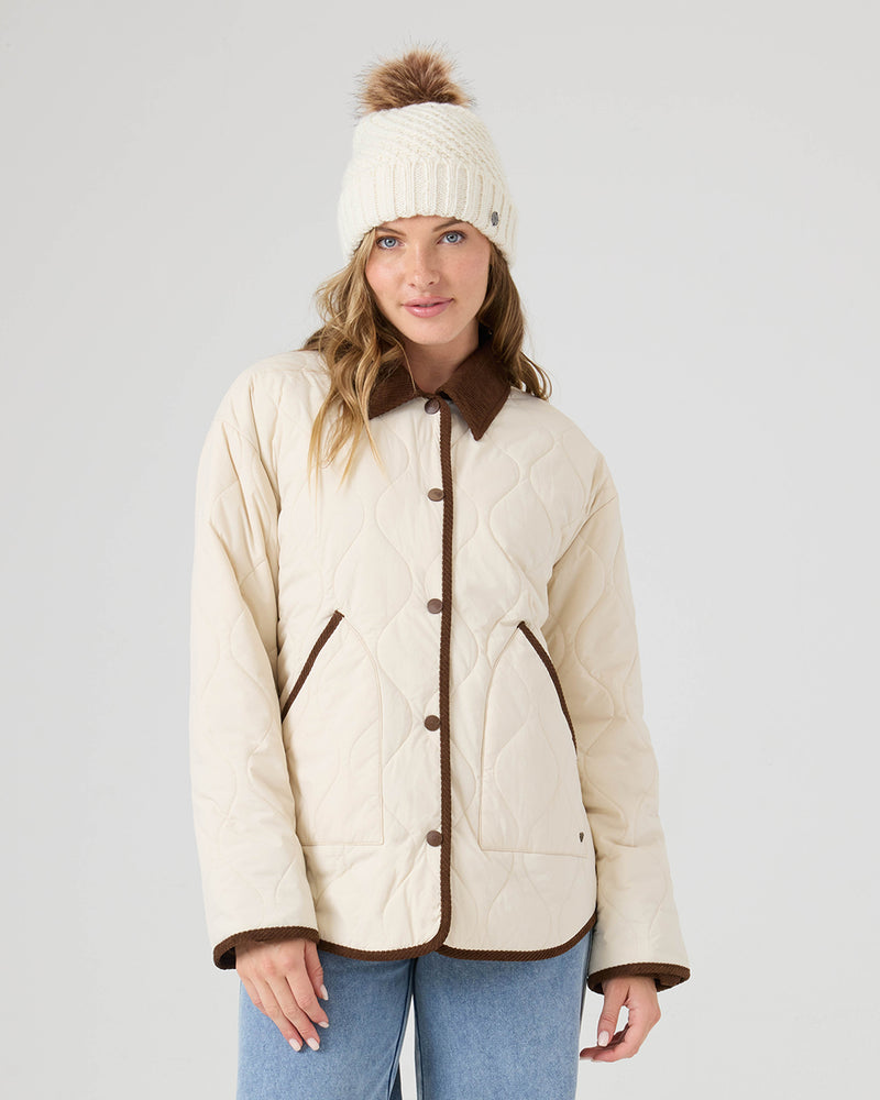 A woman wearing the Roxy Nyvea Quilted Barn Jacket in Fog, along with blue jeans and a cream knit beanie with a brown pompom, stands against a plain light background.