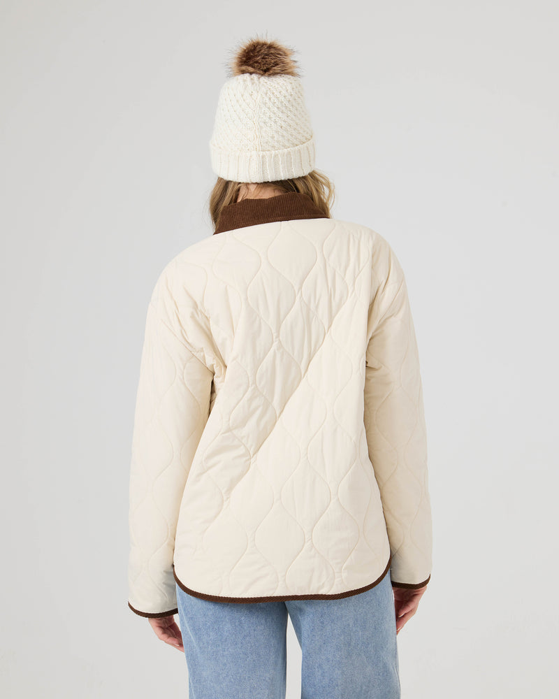 A person shown from the back against a plain white background wears the Roxy Nyvea Quilted Barn Jacket in cream, styled with blue jeans and a white knit hat featuring a brown pom-pom.