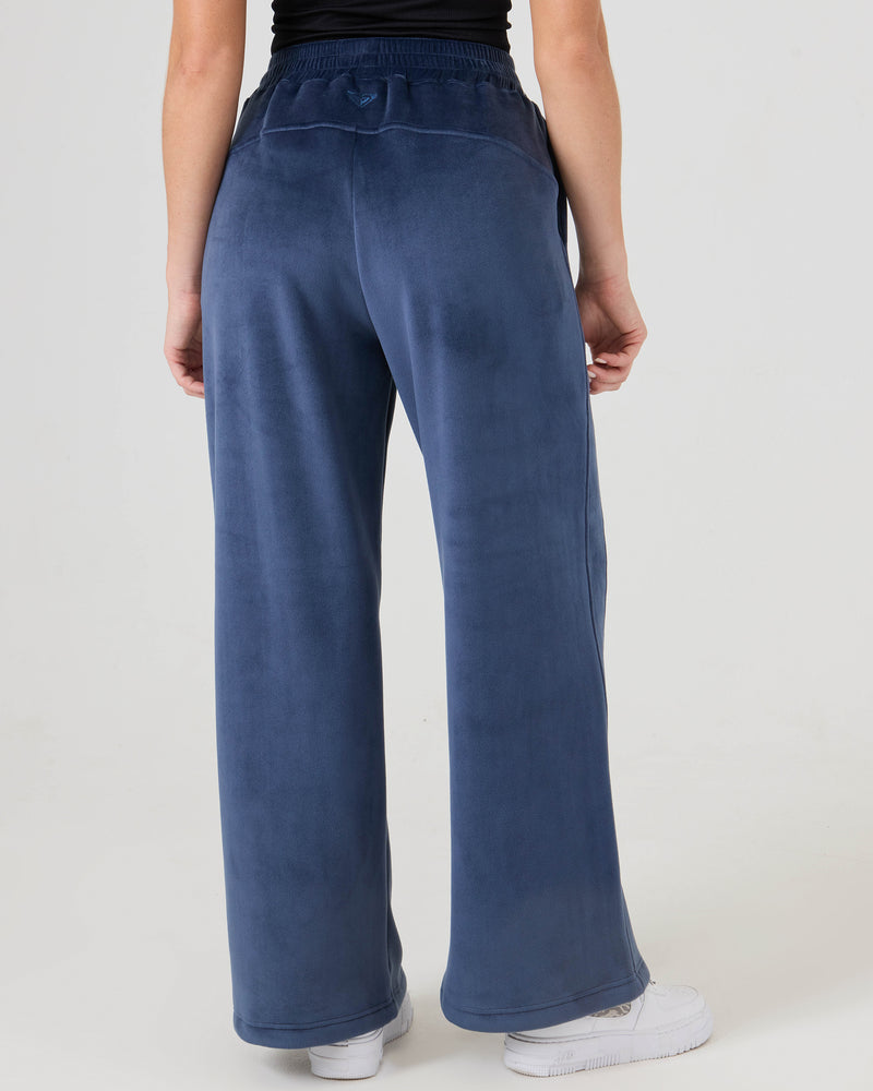 A person wearing Roxy Vivi Bonded Velour Pants and white sneakers stands with their back to the camera against a plain light background.