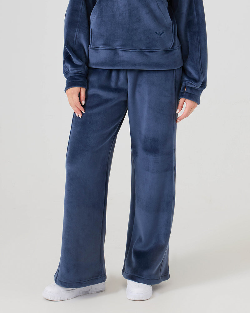 Wearing the Roxy Vivi Bonded Velour Pants in blue with a high waist and wide legs, paired with white sneakers; shown partially against a plain light background.