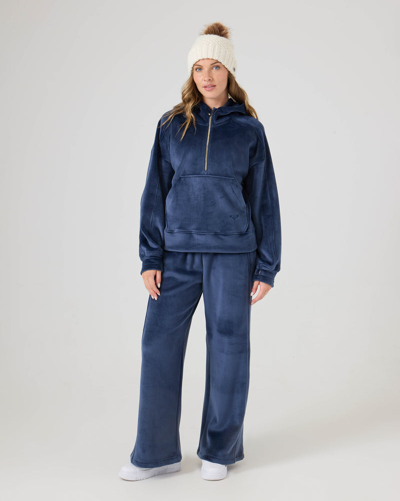 A woman wears Roxy Vivi Bonded Velour Pants with a zip-up hoodie, white sneakers, and a white knit pom-pom beanie, standing against a plain light background.
