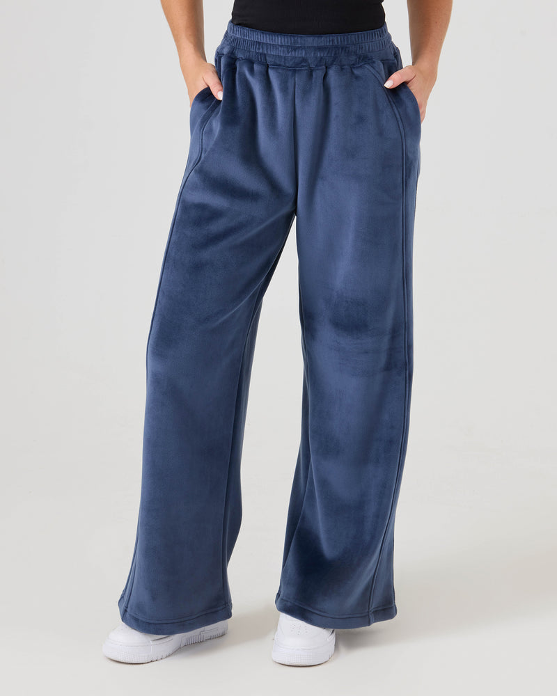 Person wearing Roxy Vivi Bonded Velour Pants in Mood Indigo, high-waisted and wide-leg with hands in pockets, styled with a black top and white shoes against a plain light background.