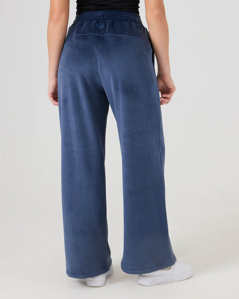 Person seen from behind wearing Roxy Vivi Bonded Velour Pants in blue, paired with a black top and white sneakers against a plain light background.