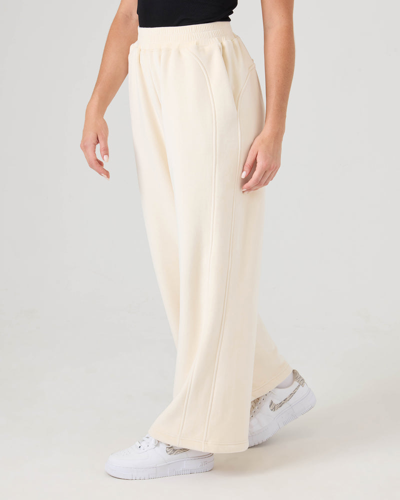 A person wearing Roxy Vivi Bonded Velour Pants in cream, a black top, and white sneakers stands against a plain light background.