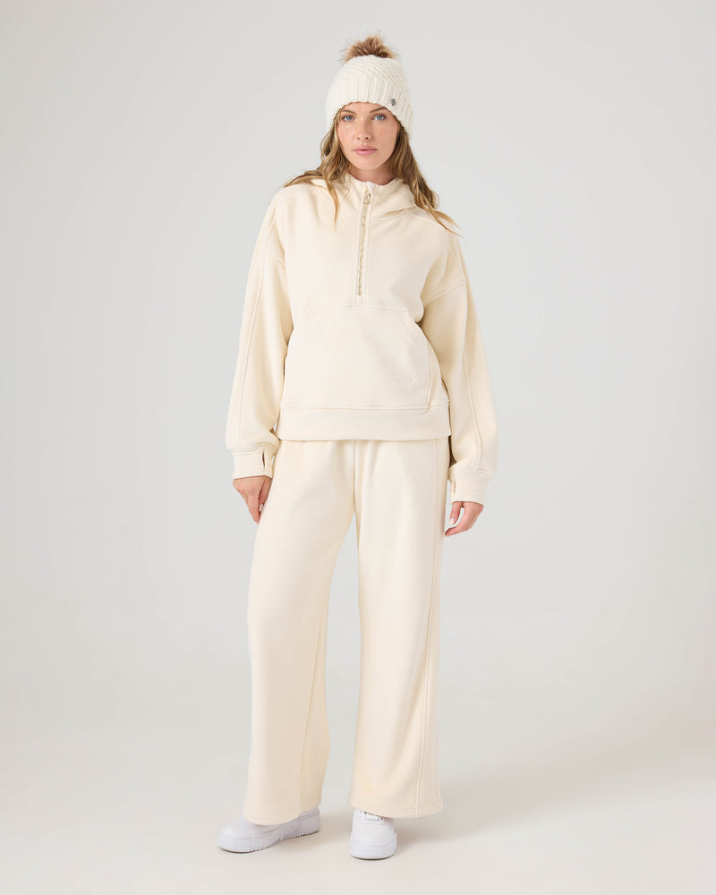 A person wearing Vivi Bonded Velour Pants by Roxy, a cream-colored hoodie, white sneakers, and a knit hat with a pom-pom stands against a plain light background.