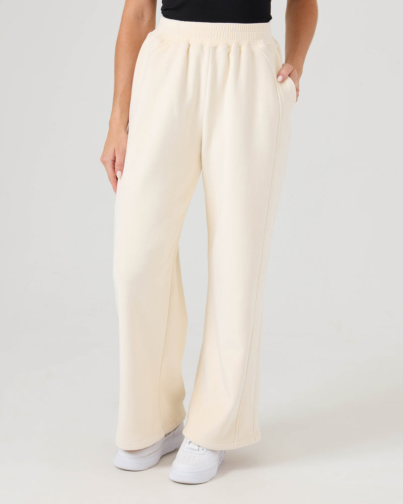 A person wears Vivi Bonded Velour Pants by Roxy in Whitecap, paired with a black top and white sneakers, standing with one hand in their pocket against a plain background.