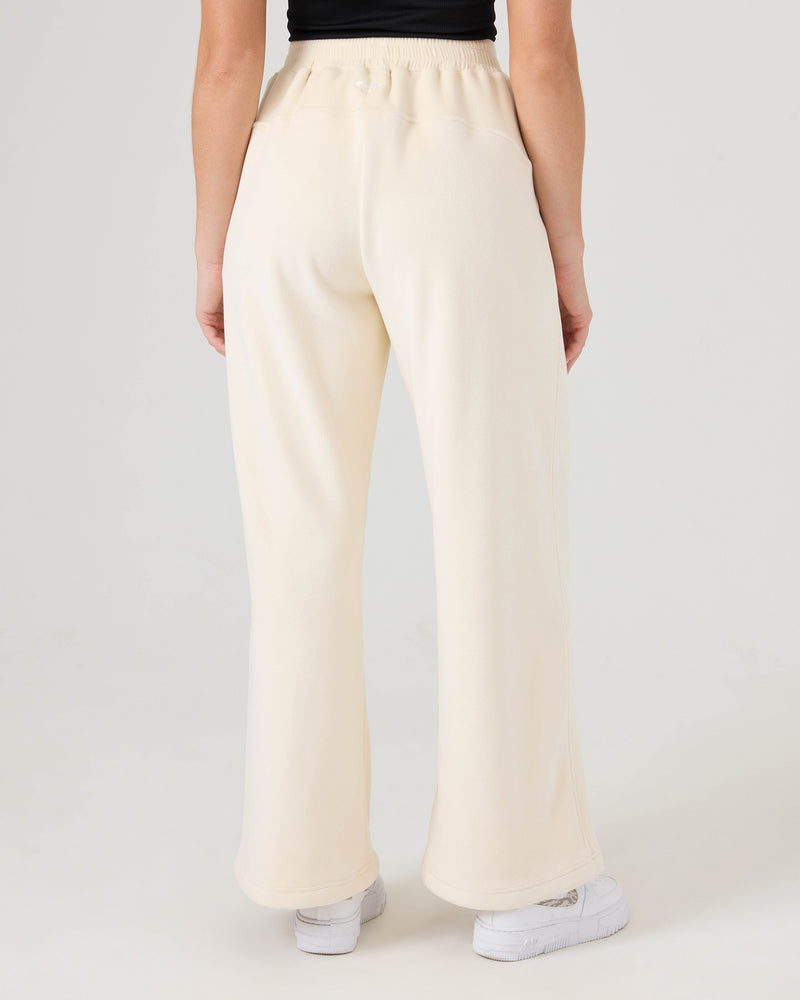 Model wears Roxy Vivi Bonded Velour Pants in cream, styled high-waisted and wide-leg with white sneakers, viewed from the back against a plain light backdrop.