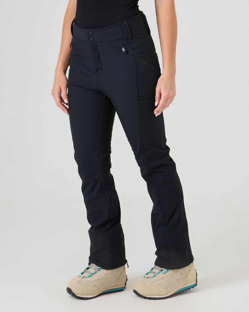 A person models the Roxy Haylee Softshell Pants—black, fitted flare pants with zippered pockets—against a plain light background. The beige shoes they wear feature teal accents, and only the lower body is visible.
