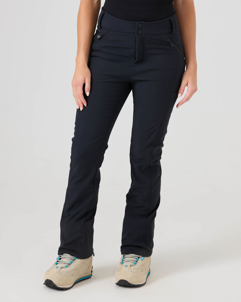 A person wearing Roxy Haylee Softshell Pants in black—fitted flare style with zippered storage—paired with a black top and beige sneakers featuring teal accents, standing against a plain light background. Only the lower half of the body is visible.