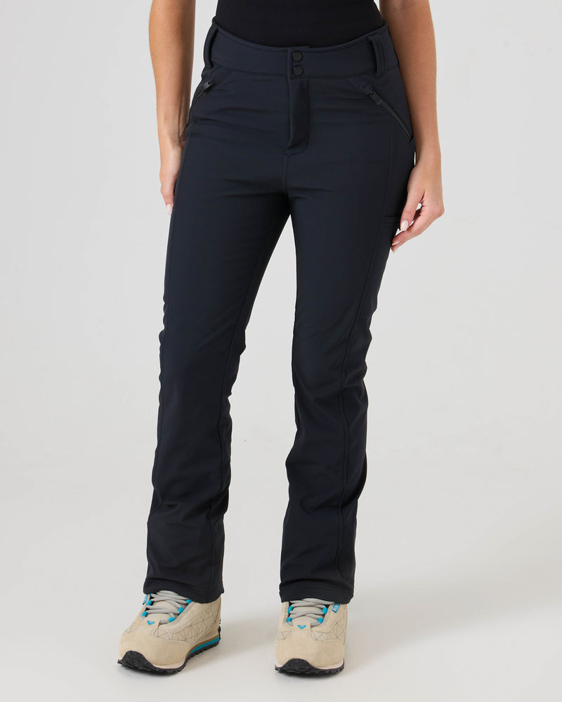 A person wearing Roxy Haylee Softshell Pants in black, paired with light tan hiking shoes featuring blue laces, stands against a plain white background. The image focuses on the lower half of the body, showcasing the sleek fit and zippered storage details of the pants.