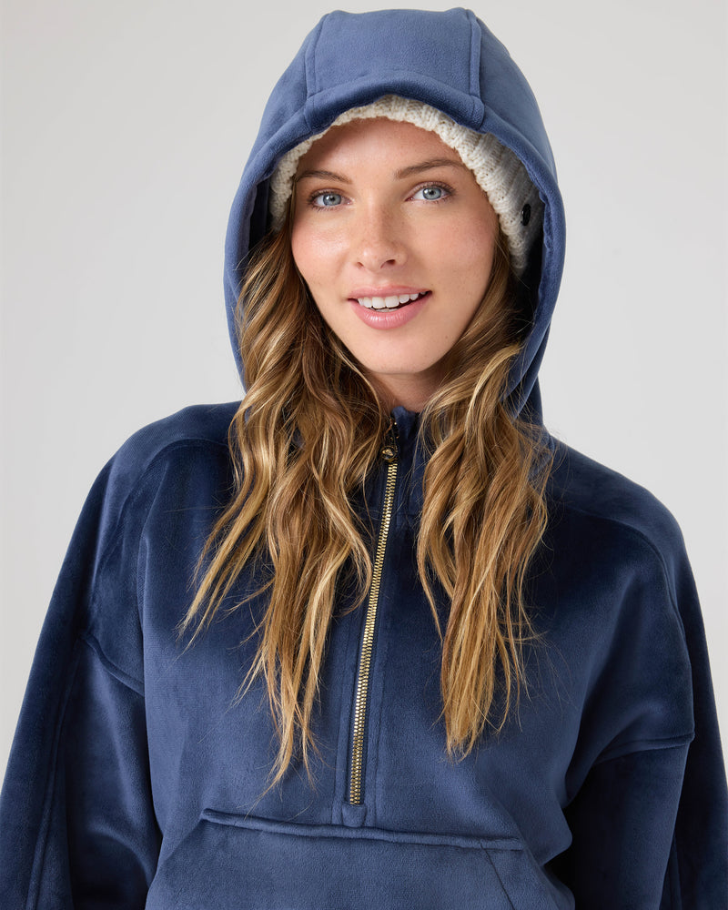 A woman in a blue Vivi Bonded Pullover by Roxy and a white knit beanie smiles at the camera, her hood up, highlighting the cozy half-zip style.