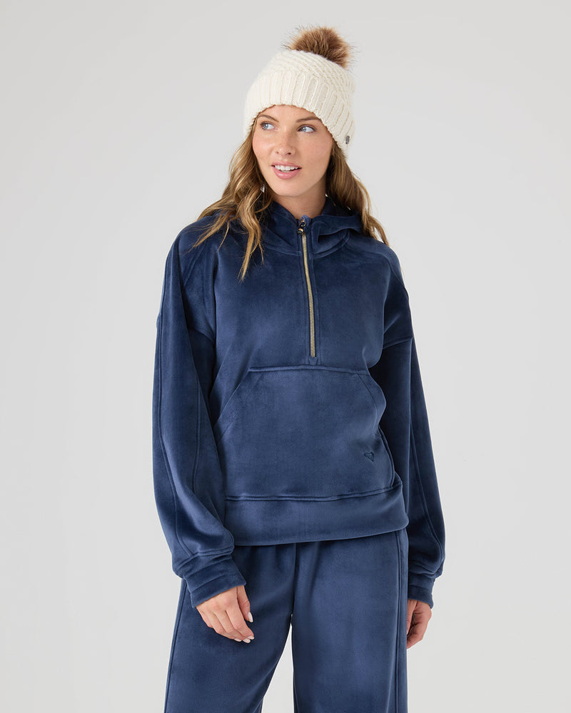 A woman in a navy blue Roxy Vivi Bonded Pullover and a white knit beanie with brown pom-pom stands slightly turned left, smiling with relaxed hands against a plain light background.
