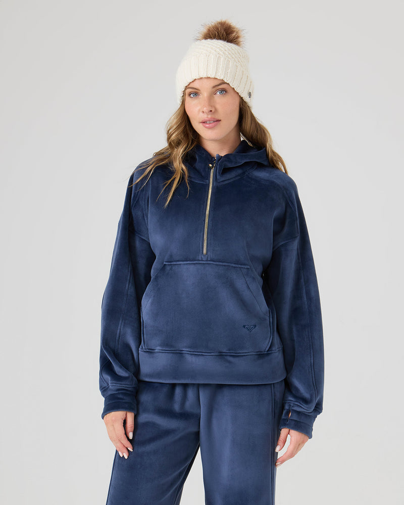 A woman in a cream knit pom-pom beanie and matching pants wears the Roxy Vivi Bonded Pullover in Mood Indigo, standing against a plain light gray background.