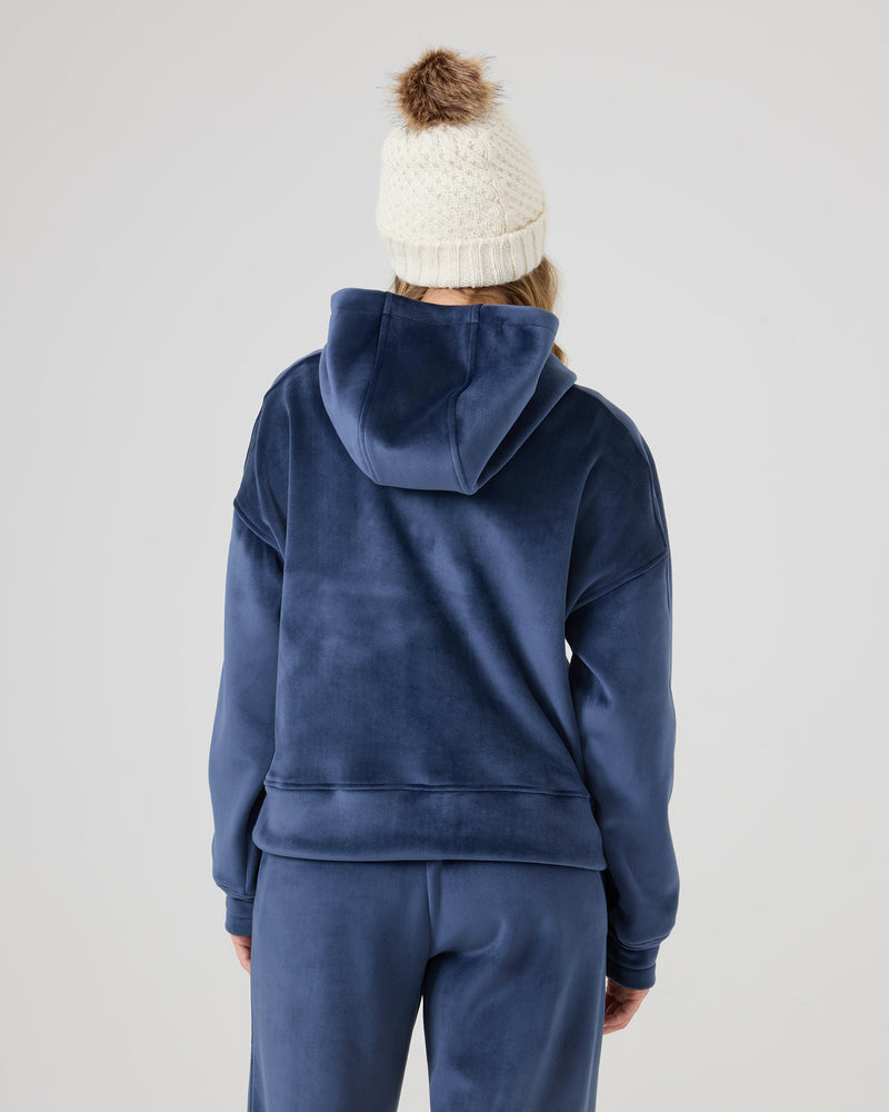 A person is shown from the back wearing the Roxy Vivi Bonded Pullover in blue velour with a white knit beanie featuring a brown pom-pom, set against a plain light background.