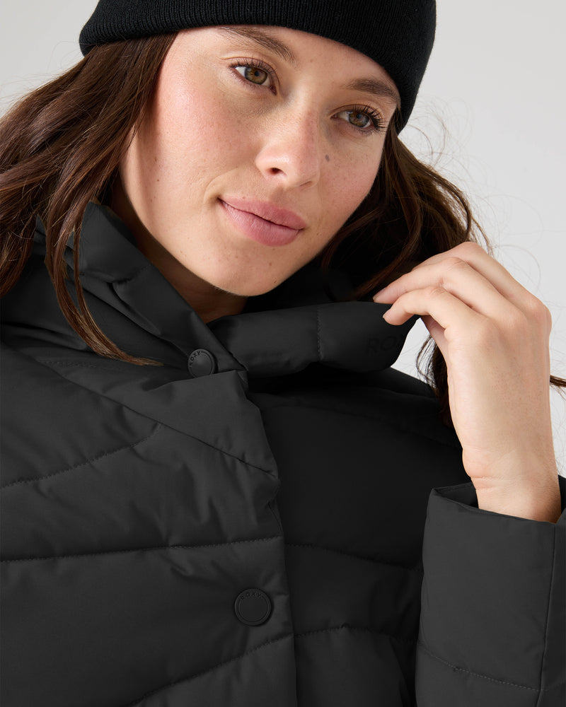 A person in a black beanie and the Abby Quilted Puffer by Roxy looks to the side, gently holding the jacket’s collar—perfect for cool-weather style.