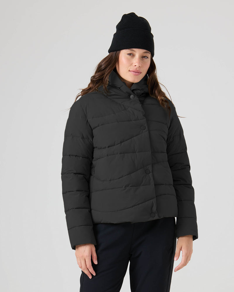 Wearing the Roxy Abby Quilted Puffer in black, a person stands against a plain light background, paired with a black beanie and pants, looking at the camera with a neutral expression.