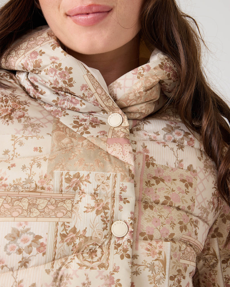 A woman wears the Roxy Abby Quilted Puffer, featuring a beige and pink floral patchwork design. The jacket is buttoned up, showing only her hair and the lower part of her face. Perfect for cool weather.