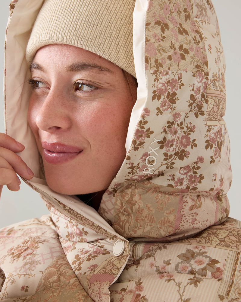 A person in a beige beanie wears the Roxy Abby Quilted Puffer, a regular fit jacket with pink and brown floral designs and a white snap button, smiling softly while looking to the left.