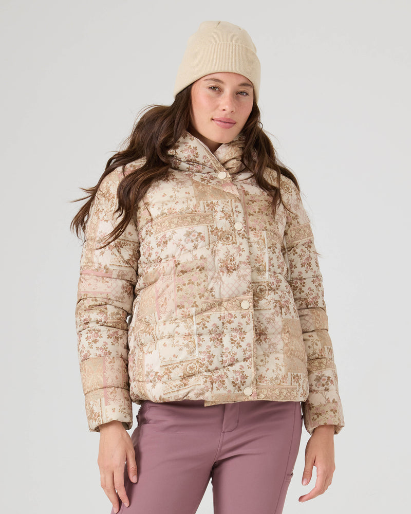 A woman in a beige knit beanie, regular fit mauve pants, and the Roxy Abby Quilted Puffer in Earthy Wahine stands against a light gray background, looking at the camera with a relaxed expression.
