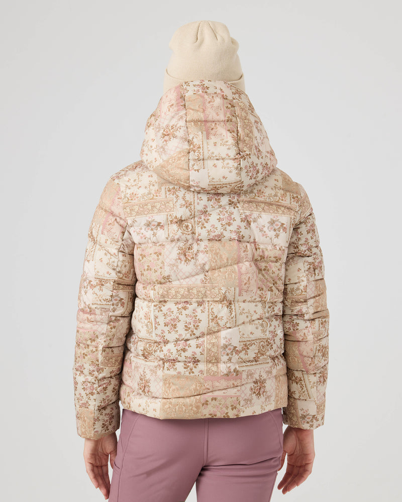 Person seen from behind in the Abby Quilted Puffer by Roxy—a floral-patterned, hooded jacket paired with mauve pants and a beige beanie. The plain, light background highlights this cool-weather look.