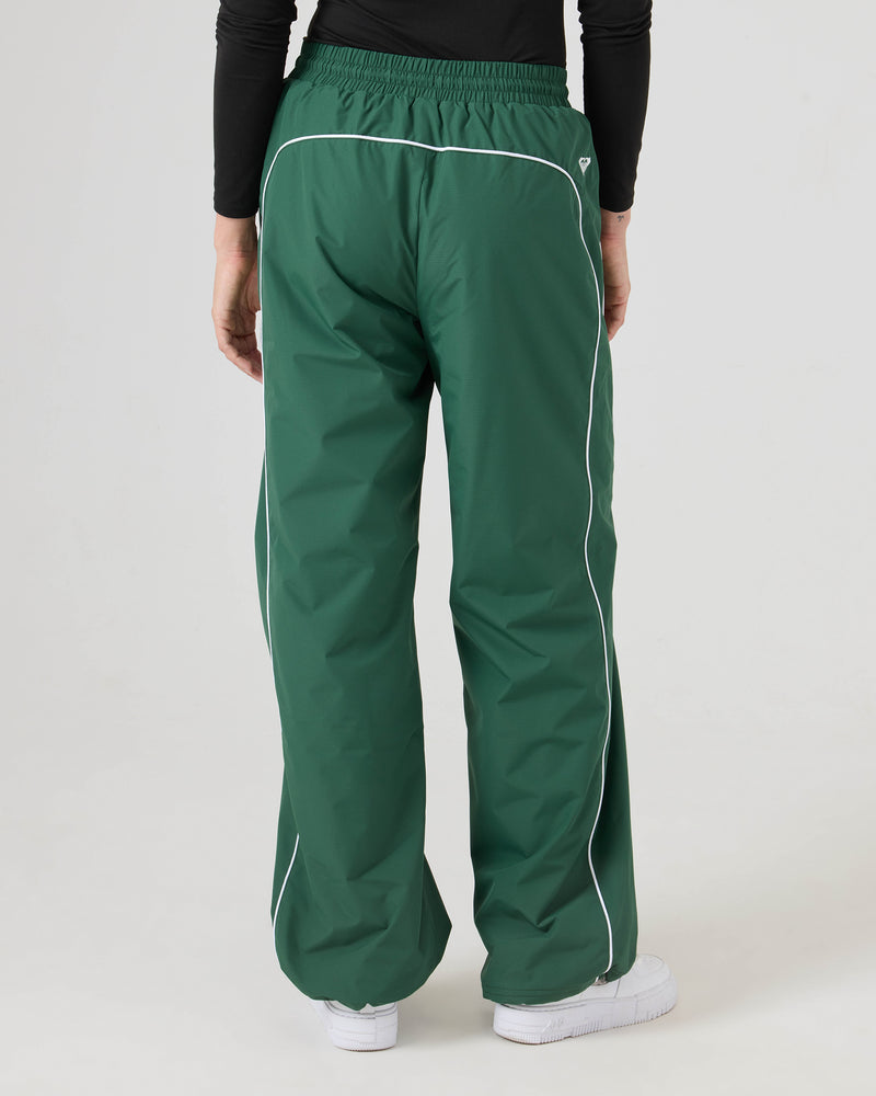 Model wears loose green Stevie Parachute Pants by Roxy with white piping, a black long-sleeve top, and white sneakers, standing against a plain light background, pictured from the back.