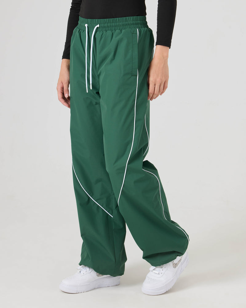 A person wears Roxy Stevie Parachute Pants—green wide-leg with white piping and adjustable waistband—paired with a black long-sleeve top and white sneakers against a plain light background.