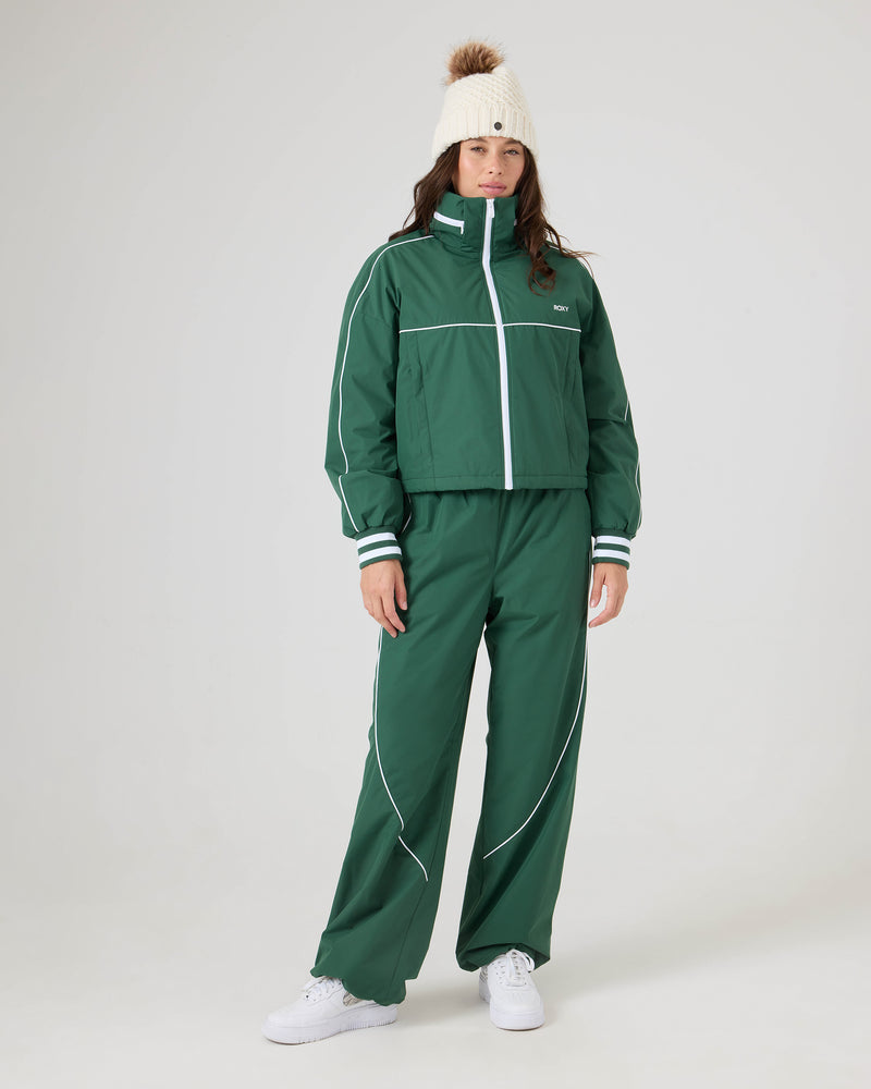A person wears Roxy Stevie Parachute Pants in green with white piping and an adjustable waistband, paired with a white pom-pom beanie and white sneakers against a plain light background.
