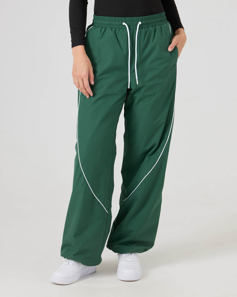 A person wears Roxy Stevie Parachute Pants in dkgreen, paired with a black long-sleeve top and white sneakers, standing against a plain light background.