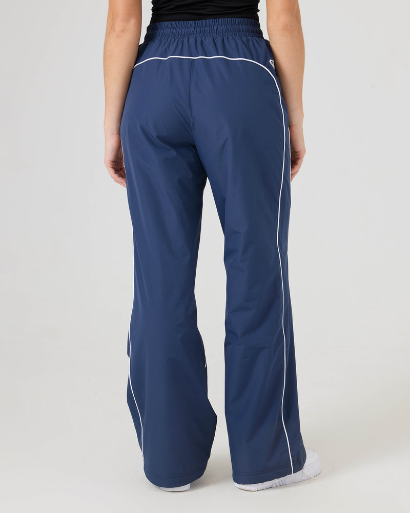 A person wearing Roxy Stevie Parachute Pants in navy blue with white piping, seen from the back. The pants have an adjustable waistband and are styled with a black top and white shoes.