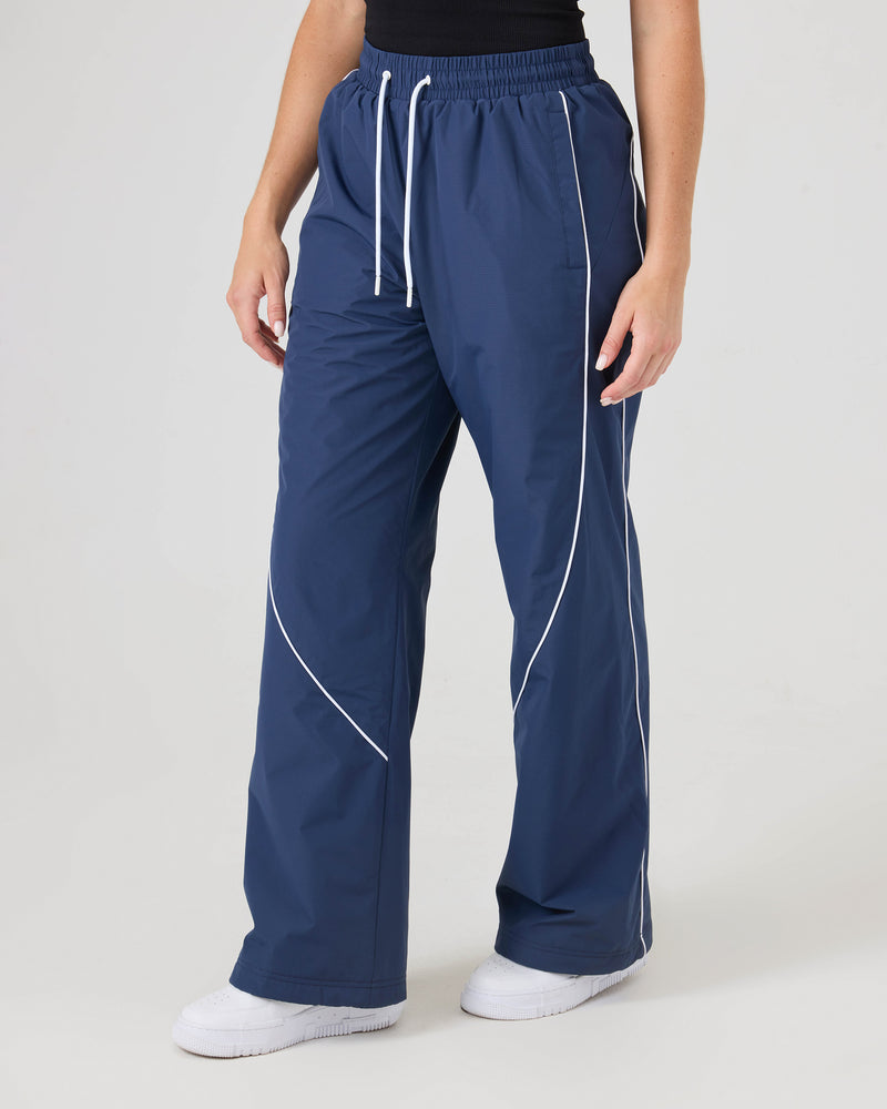 Model wears Roxy Stevie Parachute Pants in navy with white piping, paired with a black top and white sneakers. Image shows only the lower half of the body against a plain background.