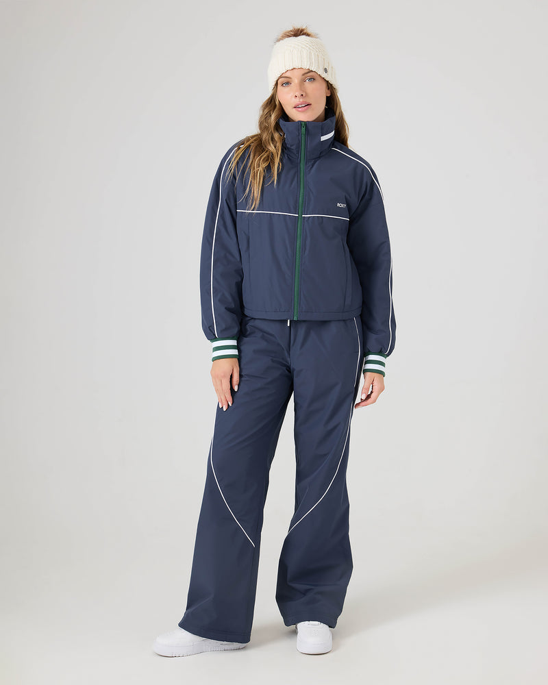 Woman in a blue tracksuit featuring Roxy Stevie Parachute Pants with white piping, wide-leg design, and adjustable waistband, paired with white sneakers and a white knit headband, facing forward against a plain light background.