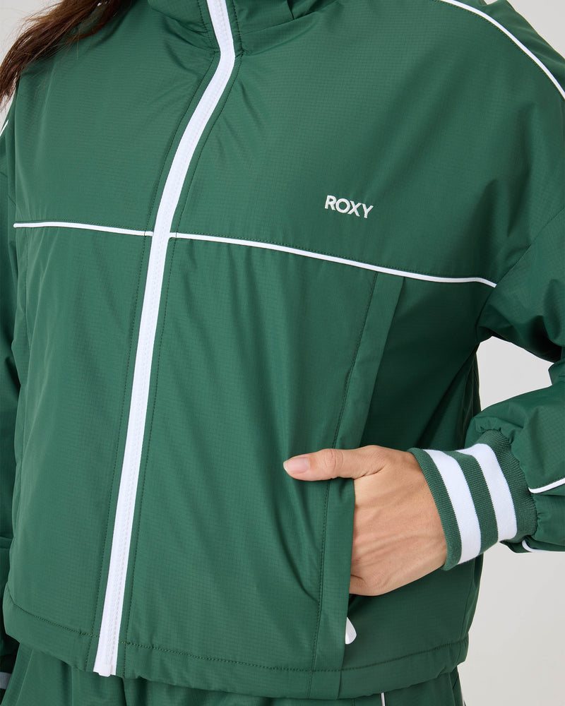 Person wearing the Emily Track Jacket by Roxy—a green retro jacket with white piping, striped cuffs, and ROXY logo. Relaxed fit, paired with matching pants; photo features torso and arm, not full face.