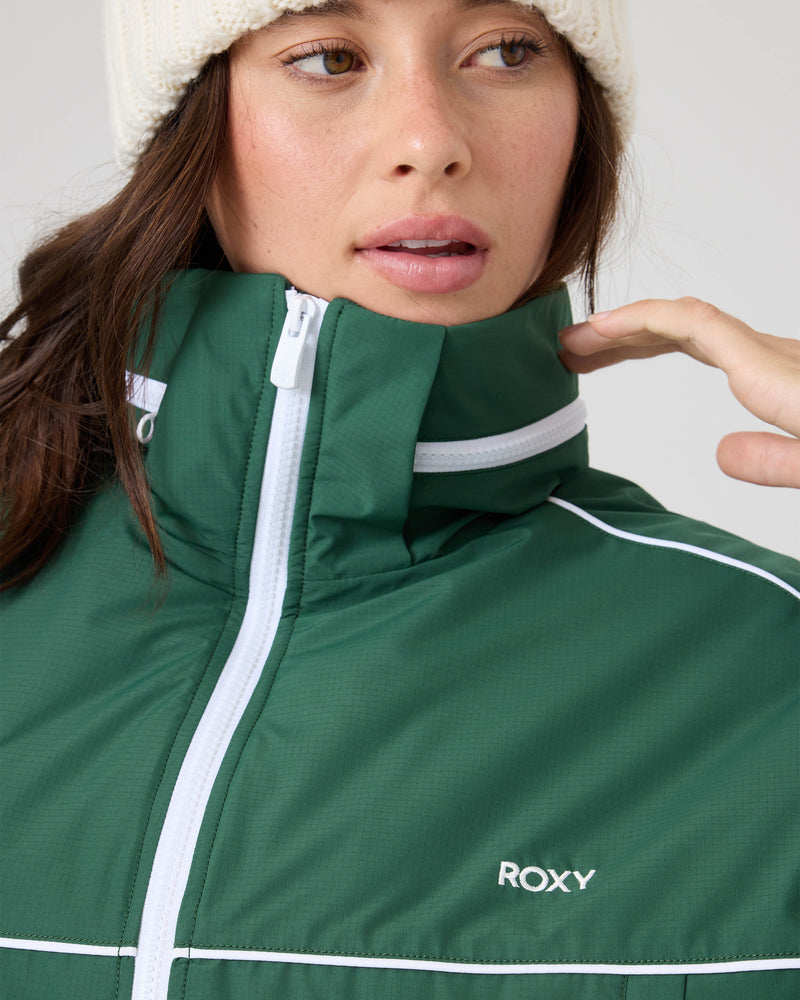 A person in the Emily Track Jacket by Roxy—a green retro style with white trim and logo—wears a cream knit beanie and looks to the side, one hand near the collar.