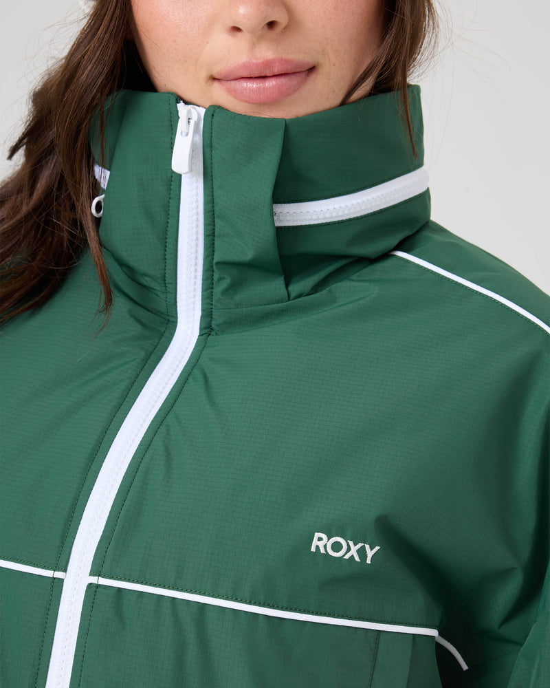 A person wears a green Emily Track Jacket by Roxy, featuring white zippers, piping, and ROXY embroidery on the chest. Only the lower face and upper torso are shown.