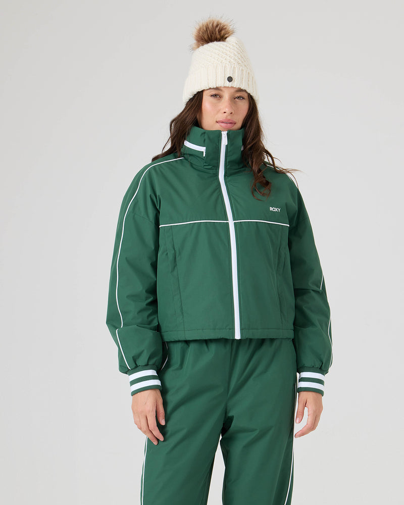 A person in the Roxy Emily Track Jacket in Dkgreen stands against a plain light background, paired with matching pants and a cream knit beanie with a brown pom-pom.