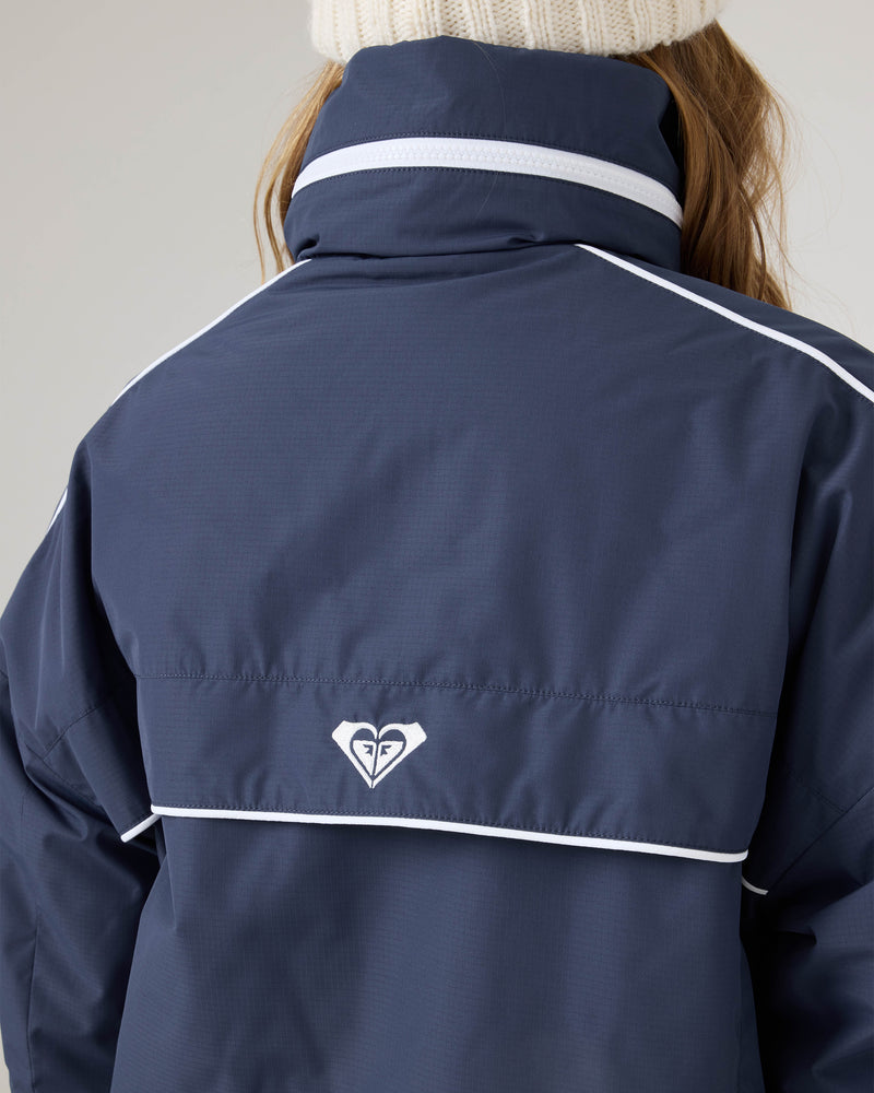 Wearing the Roxy Emily Track Jacket in navy blue with white trim and logo on the back, paired with a white knit beanie, their brown hair shows beneath the relaxed fit.