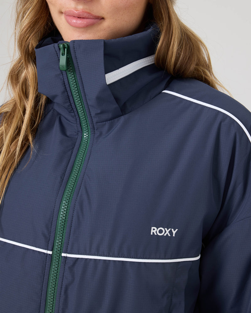 Close-up of a person wearing the Roxy Emily Track Jacket in navy blue with white piping, green zipper, and adjustable hem. Only their lower face and upper body are visible.