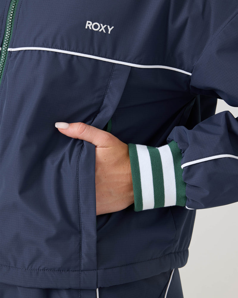 A person wearing a navy blue Emily Track Jacket by Roxy, featuring white piping and green-striped cuffs, has their hand in the jacket pocket.