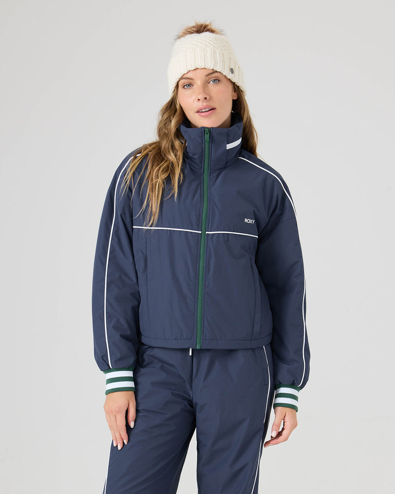 Woman wears the Roxy Emily Track Jacket in Mood Indigo, featuring white stripes and green-striped cuffs, paired with matching pants and a white knit beanie against a plain light background.