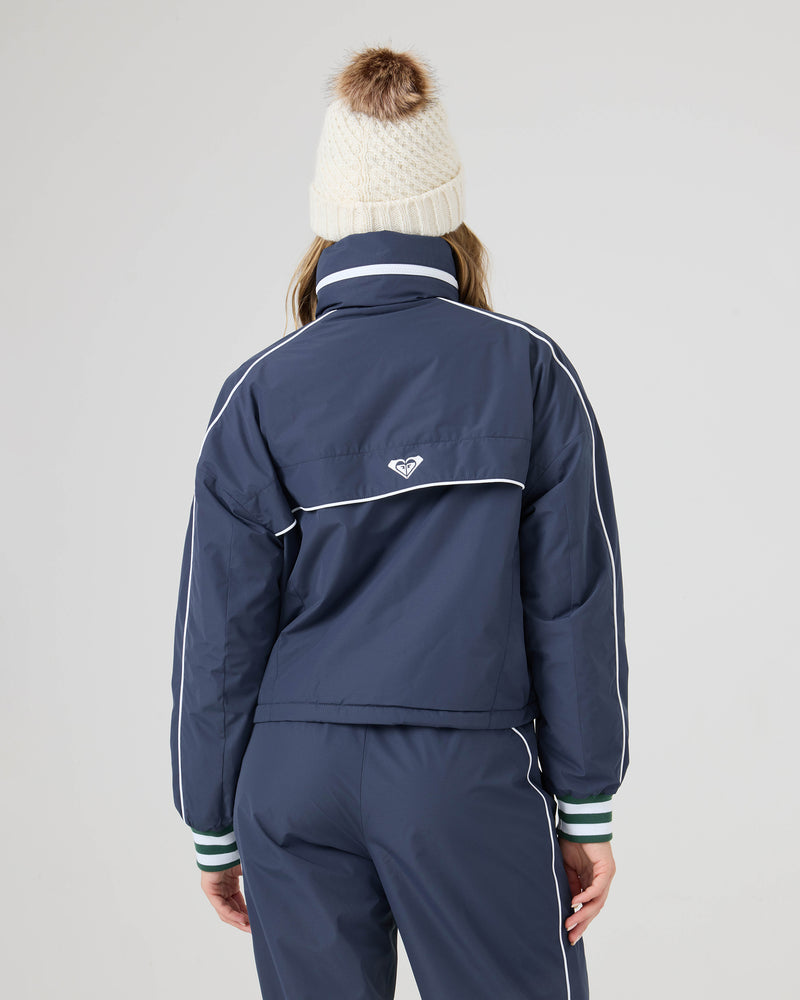 A person wears the Roxy Emily Track Jacket with white piping and a cream knit beanie with a brown pom-pom, facing away from the camera against a plain background.