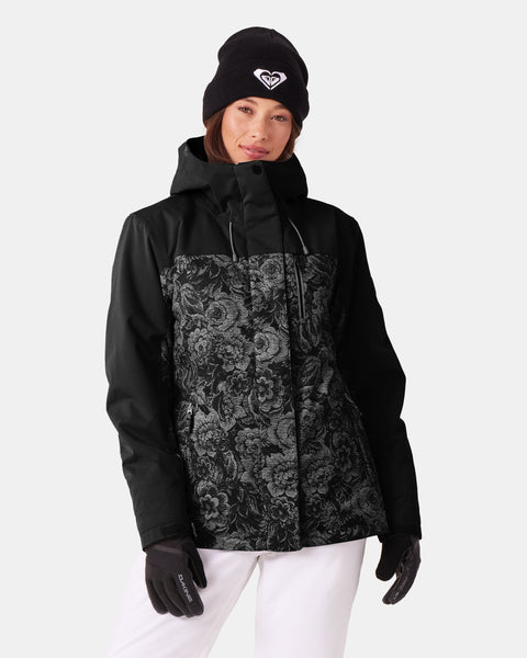 Wearing the Roxy Jetty 3N1 Jacket—a black women’s snowboard jacket with a floral pattern made from recycled materials—along with black gloves, a black beanie featuring a white logo, and white pants, a person stands against a plain white background.