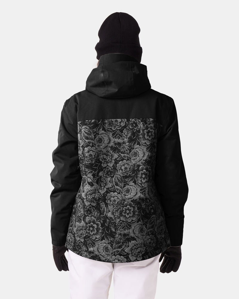 A person wearing a black beanie and gloves is shown from behind against a plain light background. They are wearing the Roxy Jetty 3N1 Jacket—a women’s snowboard jacket with a 3-in-1 design, featuring a black exterior with a floral pattern on the back—paired with white pants.