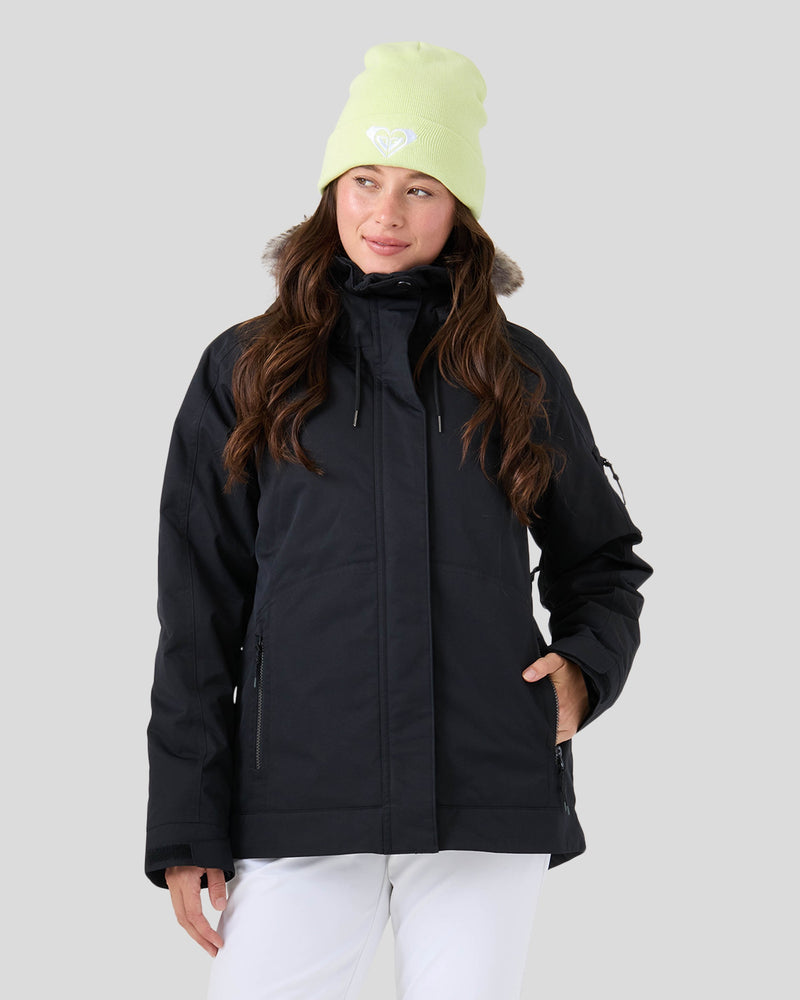 A woman stands against a plain light gray background wearing the Roxy Meade Jacket—a black winter coat with a fur-trimmed hood designed for insulated warmth—paired with white pants and a light yellow beanie featuring a white heart logo.
