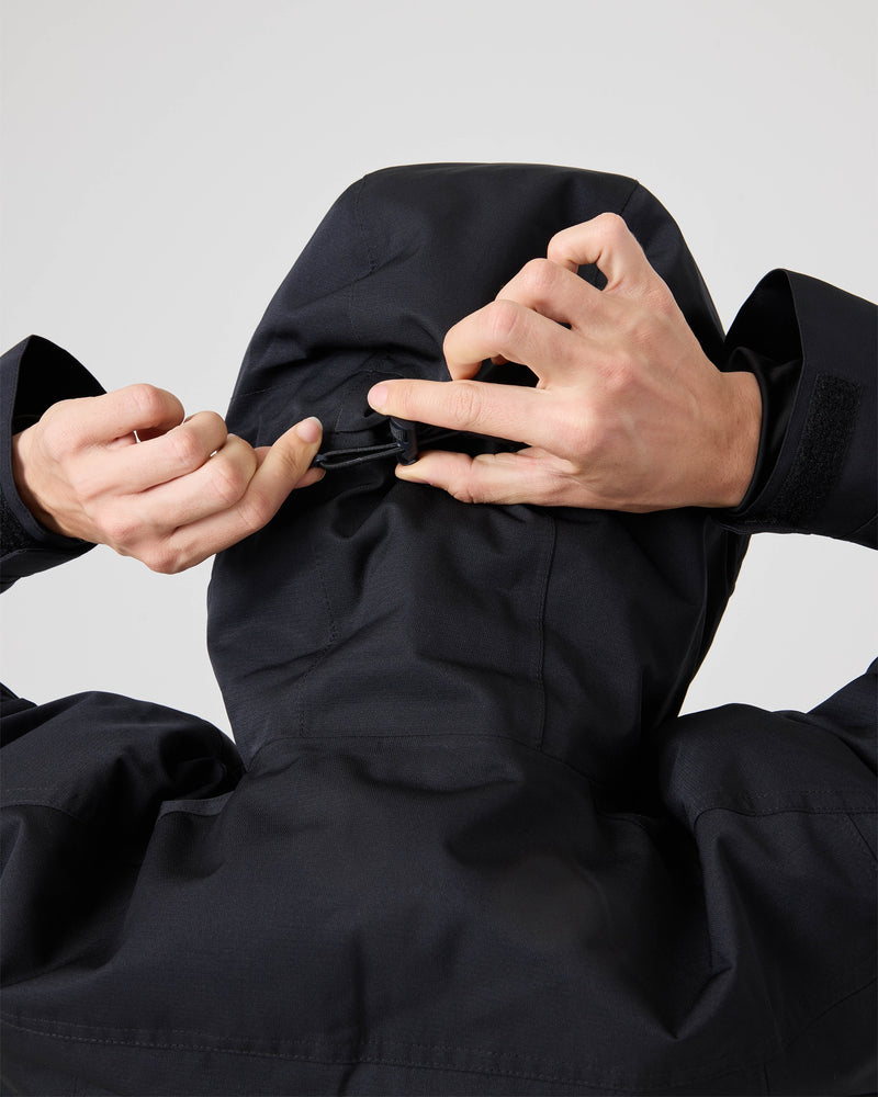 A person adjusts the drawstring at the back of a dark Roxy Meade Jacket, with both hands visible. The image is captured from behind against a plain, light background. This women’s snowboard jacket provides insulated warmth and waterproof protection for winter conditions.
