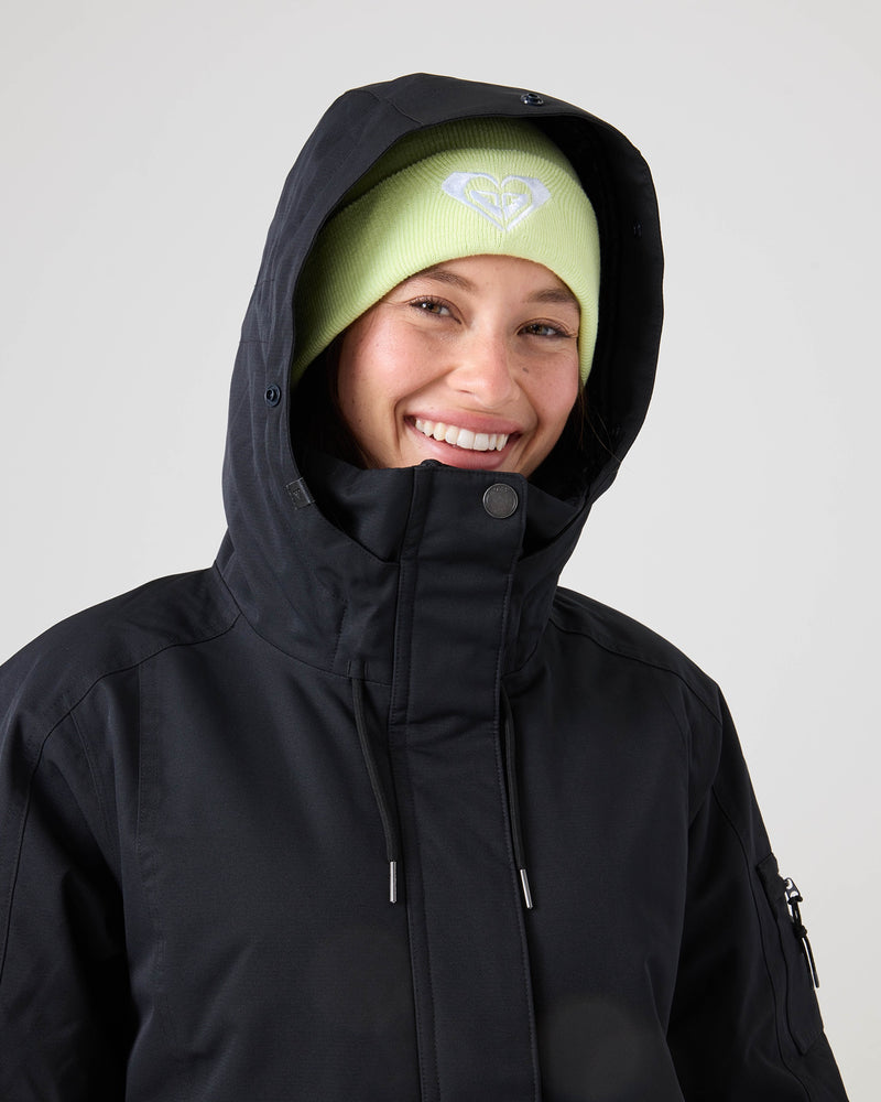 A person smiles against a plain light background, wearing the Roxy Meade Jacket—a black hooded coat designed for insulated warmth and waterproof protection—and a light green beanie adorned with a heart-shaped logo.