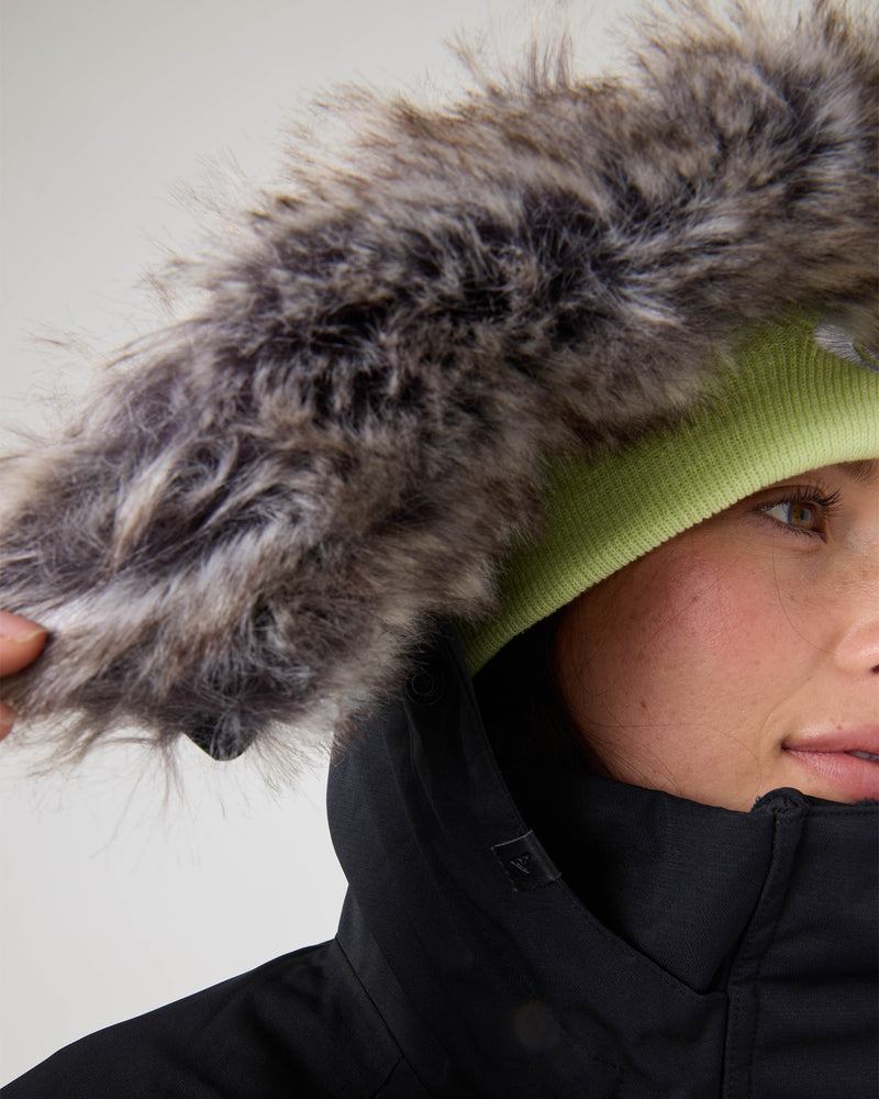 Close-up of a person wearing a lime green beanie and black coat with a furry hood, highlighting the insulated warmth and waterproof protection of the Roxy Meade Jacket. Only part of their face is visible as they hold the edge of the hood.