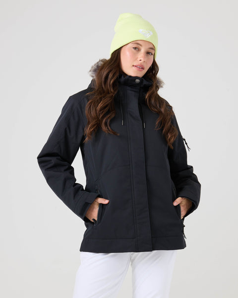 A woman wears a light green beanie and white pants, standing with her hands in her pockets as she models the Roxy Meade Jacket—a black women’s snowboard jacket with a fur-trimmed hood—against a plain light background, offering both insulated warmth and style.