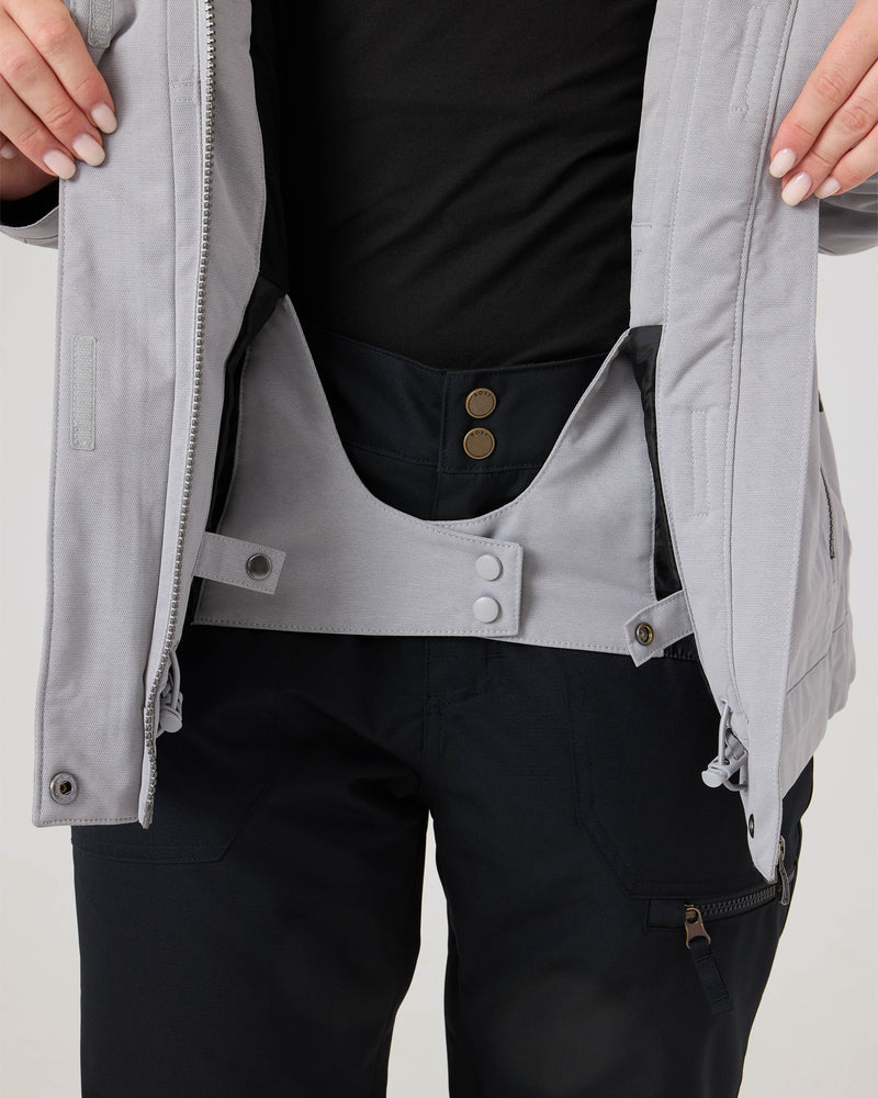 A person wearing black pants and a black shirt demonstrates fastening the inner snow skirt of the light gray Roxy Meade Jacket for women using snap buttons, highlighting its insulated warmth ideal for chilly days.