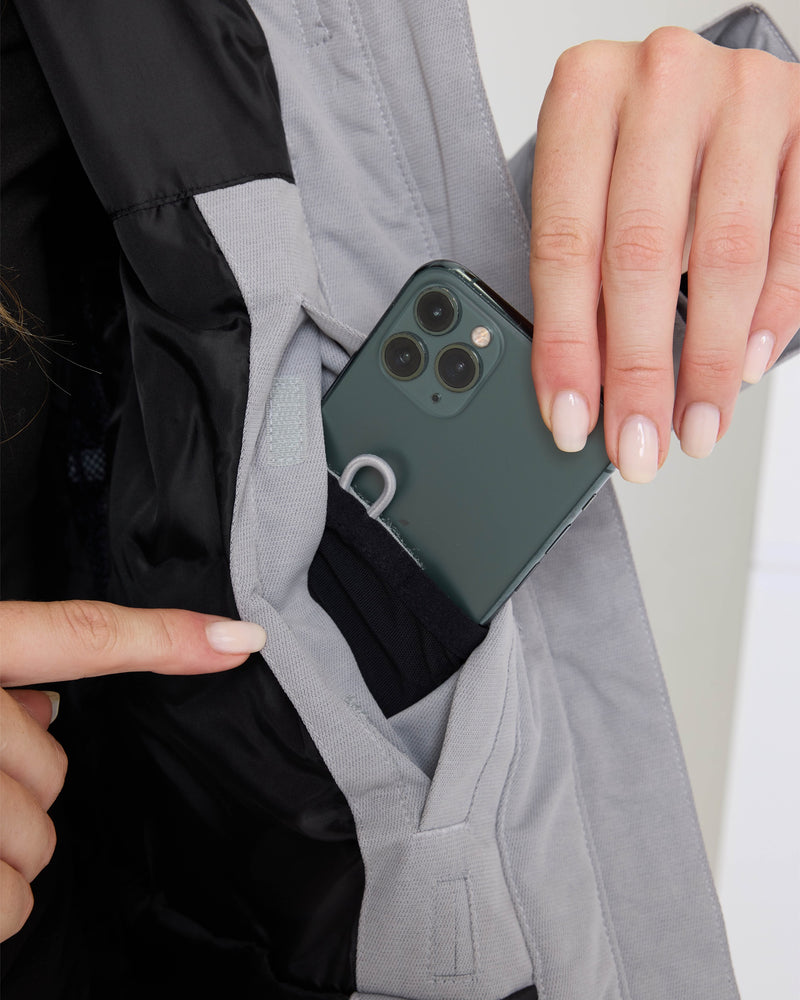A person places a smartphone into the inner pocket of a light gray Roxy Meade Jacket, using one hand to hold the phone and the other to open the pocket.